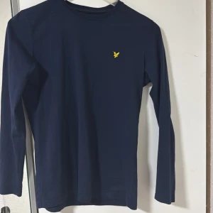 Marinblå långärmad tröja Lyle & Scott - Snygg marinblå långärmad tröja från Lyle & Scott Junior med gul logga på bröstet. Tillverkad i 100% bomull för skön känsla och passform. 