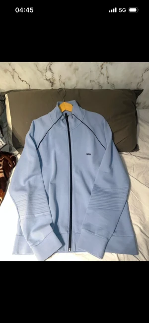 Ljusblå ziptröja från BOSS XXL - Snygg ljusblå ziptröja från BOSS i storlek XXL. Tröjan har hel dragkedja, ståkrage och diskret BOSS-logga på bröstet. Materialet är mjukt och känns som bomull/polyester. Snygga detaljer med svarta sömmar och ribbade partier på ärmarna.