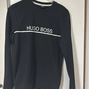Svart långärmad tröja Hugo Boss - Svart långärmad tröja från Hugo Boss med vit logotyp och linje över bröstet. Rund hals med vit detalj på insidan av kragen. Storlek S men passar M 