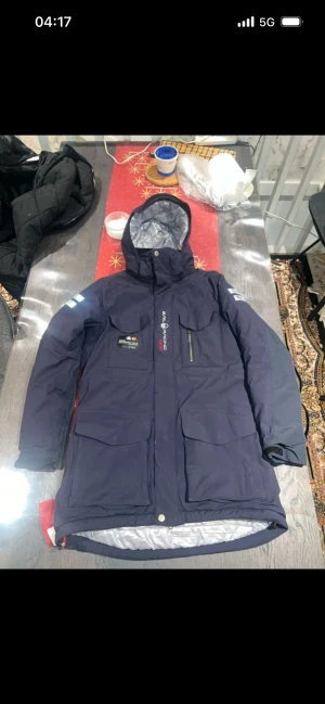 Sail Racing blå dunjacka med huva - Blå dunjacka från Sail Racing med huva, stora fickor och reflekterande detaljer på ärmarna. Insidan är quiltad i silver och svart, och jackan har dragkedja samt knappar framtill. Perfekt för kalla dagar och har en snygg, sportig look.