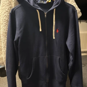 Mörkblå hoodie från Polo Ralph Lauren - Snygg mörkblå hoodie från Polo Ralph Lauren med dragkedja och huva. Tröjan har vita snören, två fickor fram och rött broderat Polo-logo på bröstet. HELT NY säljs för att den inte passat och gått förlångtid för att skicka tillbaka