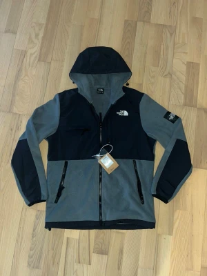 The North Face grå och svart fleecejacka - Grym grå och svart fleecejacka från The North Face med huva och dragkedja. Jackan har flera fickor med dragkedja och logga på bröstet och ärmen. Perfekt för kyliga dagar och outdoor vibes.