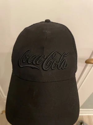Svart Coca-Cola keps med mesh - Svart keps med broderad Coca-Cola-logga framtill. Framsidan är i tyg och baksidan har mesh-paneler för extra ventilation. Justerbar snapback baktill. Snygg och stilren design som passar till streetwear och casual outfits.