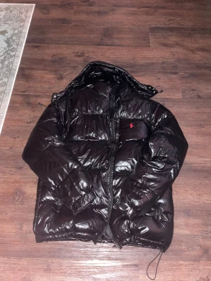 Svart pufferjacka från Polo Ralph Lauren - Säljer en svart, glansig pufferjacka från Polo Ralph Lauren med huva och dragkedja framtill. Jackan har det klassiska röda Polo-logot på bröstet och är riktigt bra för kalla dagar och dunfylld för extra värme.