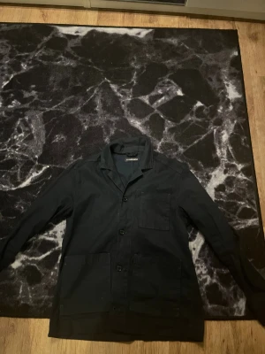 Mörkblå overshirt från J.Lindeberg - Snygg mörkblå overshirt från J.Lindeberg med klassisk krage och tre stora fickor framtill. Jackan har knäppning med svarta knappar och är tillverkad i bomull. Perfekt för dig som gillar stilrena och enkla plagg med lite workwear-vibe.