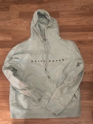 Mintfärgad Daily Paper hoodie - Säljer en ljusblå hoodie från Daily Paper med svart text på bröstet. Storlek M. Små ljusa flöckar på backsidans ena axel men syns knappt.