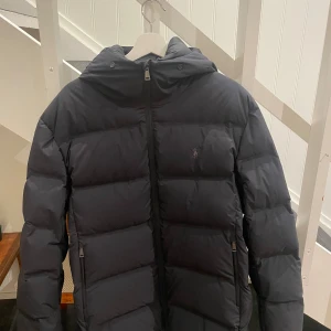 Svart pufferjacka från Polo Ralph Lauren - Svart pufferjacka från Polo Ralph Lauren med Pertex Unlimited-tyg. Jackan har huva, raka quiltade paneler och dragkedja framtill. Diskret Polo-logga på bröstet och två sidofickor med dragkedja. Perfekt för kalla dagar med stil. Det är storlek M men är stor i storlek. Oanvända säljs på grund av fel storlek. Priset går att diskutera!