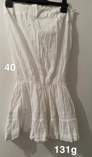 Vit ärmlös tubtopp med spetsdetaljer - Säljer en vit ärmlös tubtopp med smockad midja och fina spetsdetaljer längst ner. Toppen har en luftig och romantisk känsla med volanger och är perfekt för varma dagar. Materialet är mjukt och lätt, vilket gör den skön att bära.🌸 Fint använt skick. Storlek 40