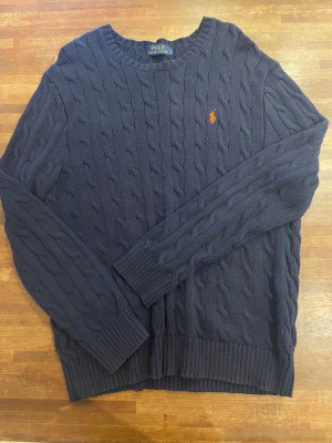Kabelstickad Polo Ralph Lauren - Snygg marinblå stickad tröja från Polo Ralph Lauren med klassiskt kabelmönster och orange logga på bröstet. Tröjan har rund halsringning, ribbade muddar och lång ärm. Perfekt för dig som gillar stilrena och tidlösa plagg.