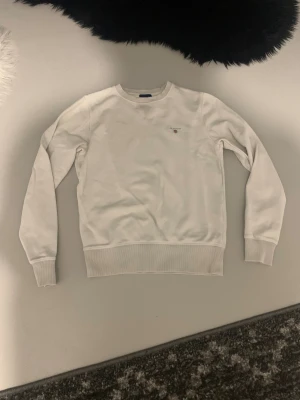 Vit sweatshirt från GANT XS - Vit sweatshirt från GANT i storlek XS. Tröjan har rund hals, ribbade muddar och ett broderat GANT-logo med sköld på bröstet. Tillverkad i mjuk bomull, perfekt för chill dagar. Enkel och stilren design som funkar till jeans eller mjukis.