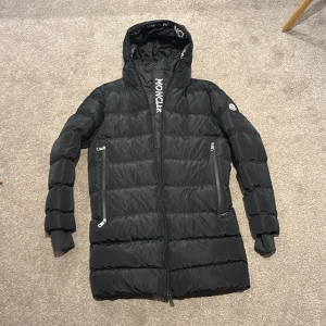 Svart Moncler dunjacka lång - Säljer en svart dunjacka från Moncler med huva och logotyp på ärmen. Jackan har quiltad design, dragkedja framtill och två stora fickor med dragkedja. Jackan har även som en diskret liten väst inuti så att man kan ha jackan öppen om man vill men ändå bli varm(se sista bild) jag är 186 och jackan passar perfekt.  Passar perfekt för kalla vinterdagar🔥🔥Skriv vid minsta fundering👍🏼. Pris kan diskuteras✅✅ är även helt öppen för byten.
