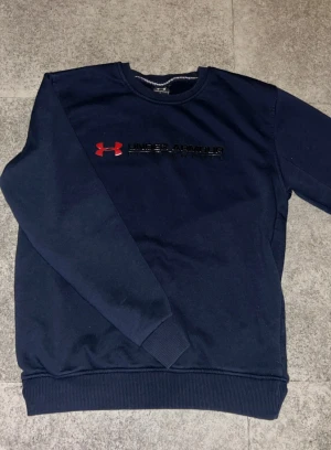 Mörkblå sweatshirt från Under Armour - Snygg mörkblå sweatshirt från Under Armour med rött och svart logotryck på bröstet. Tröjan har rund hals, ribbade muddar och är tillverkad i mjuk bomullsmix. Perfekt för chill eller träning, klassisk stil med sportig vibe.