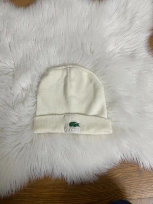 Vit mössa från Lacoste - Snygg vit mössa från Lacoste med klassisk broderad krokodil-logga framtill. Mössan är ribbstickad och har uppvikt kant för extra stilpoäng. Perfekt för dig som vill ha en clean och enkel look med en touch av street. Materialet är mjuk bomull som känns skönt mot huden.