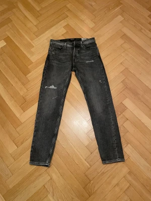 Svarta tapered jeans från Jack & Jones - Snygga svarta jeans från Jack & Jones i modellen Tapered/Mike. Jeansen har en tvättad look med slitna detaljer. Passformen är tapered vilket gör dom smalare ned mot benen. Perfekta till sneakers och hoodie. Storlek 28/32. Dessa jeans är använda vid ett fåtal tillfällen då dom inte passar mig. Bara att höra av sig vid funderingar