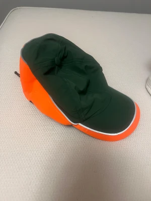 En Leif keps från Leif och Billy serien  med snörning - Säljer en unik keps i mörkgrönt och neonorange med vit kant runt skärmen. Kepsen har en böjd skärm och justerbar snörning baktill för perfekt passform. Materialet är syntet och kepsen har en cool, sportig vibe som sticker ut.