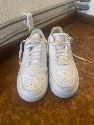 Nike Air Force 1 vita sneakers - Fräscha vita Nike Air Force 1 sneakers med klassisk låg siluett. Skorna har snörning, perforerad tå och rundad form. Tillverkade i skinn med mjukt meshfoder för extra komfort. Snyggt Nike-märke på hälen och sidan. Perfekt för dig som gillar clean och stilren look.