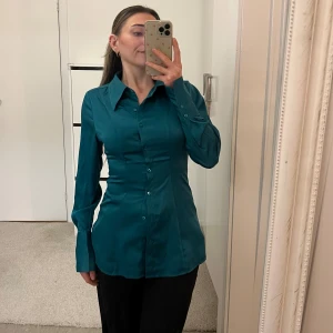 Turkos skjorta från Gina Tricot - Snygg turkos skjorta från Gina Tricot med figurnära passform, långa ärmar och klassisk krage. Skjortan har knappar hela vägen framtill och är tillverkad i ett glansigt satin material som ger en lyxig känsla. Perfekt för jul och nyår. Helt ny oanvänd, endast tvättad.