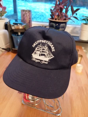 Ny keps Skeppsbyggare - Säljer en marinblå keps med vit broderad text 'Skeppsbyggare' och motiv av ett segelfartyg från Briggen Gerda. Klassisk form med böjd skärm och justerbar baksida. Perfekt för dig som gillar nautiska vibbar och snygga detaljer.