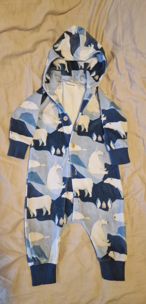 Velour jumpsuit med isbjörnar, sälar och pingviner - Supermysig jumpsuit från Ma-ia family med huva och knappar framtill. Mönstrad med vita isbjörnar, sälar och pingviner på blå bakgrund, ribbade muddar i mörkblått och tillverkad i mjuk polyester. Perfekt för kalla dagar och chill hemma.
