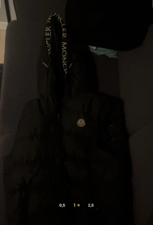 Svart Moncler väst - Svart väst från Moncler med stor huva och tydlig Moncler-logga längs kanten. Jackan har dragkedja framtill, quiltad design och klassisk Moncler-märke på bröstet. Loggan går att skanna och är en perfekt julklapp till din partner. Säljs då den inte längre används. Ser helt ny ut och finns inga fläckar eller fel på den MVH Arvid!