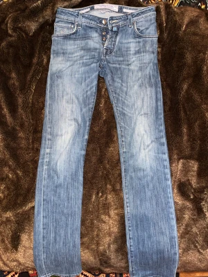 Blå jeans från Jacob Cohen Storlek 31 - Snygga blå jeans från Jacob Cohen med klassisk femficksdesign och ljus tvätt. Byxorna har raka ben, kontrastsömmar och en lila-rutig patch bak i midjan. Jeansen är tillverkade i mjukt denim och har knappgylf framtill. Storlek 31. Modell 622