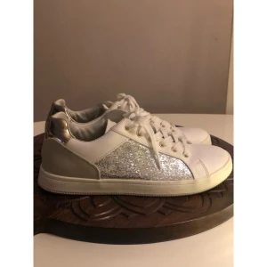 Vita sneakers med glitter och guld, 38. - Snygga vita sneakers med glittriga silverpaneler på sidorna och metallicguld-detalj på hälen. Skorna har vita snören, guldiga öljetter och en platt sula. Perfekta för dig som vill sticka ut med lite extra bling i vardagen. Märke: only. Storlek: 38