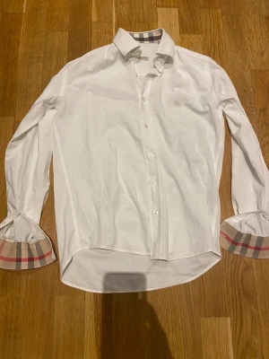Vit Burberry Brit skjorta med rutig detalj - Vit skjorta från Burberry Brit med klassisk krage och långa ärmar. Insidan av manschetterna och kragen har den ikoniska Burberry-rutiga detaljen i beige, svart och röd. Skjortan har knappar framtill och ett broderat Burberry-emblem på bröstet. Perfekt för dig som gillar stilrena och lyxiga plagg.