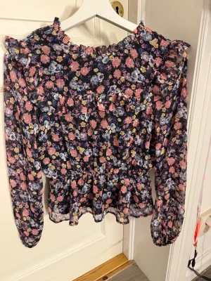Blommig volangblus från Lindex - Supersöt blommig blus från Lindex i storlek 152. Svart botten med rosa, lila, blå och gula blommor, volangdetaljer vid axlar och bröst samt lång ärm. Blusen har en lätt och luftig känsla, perfekt för dig som gillar romantisk stil.