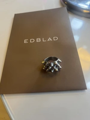 Silvrig ring med nitar från Edblad - Cool och chunky ring från Edblad i blankt silvrigt metallmaterial. Ringen har flera små nitar som ger en edgy och unik look. Perfekt för dig som gillar statement-smycken och vill sticka ut med din stil.