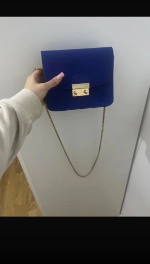 Kornblå axelväska med guldkedja - Snygg kornblå axelväska i skinnimitation med stilren fyrkantig form. Väskan har en lång guldfärgad kedja som axelrem och ett elegant guldfärgat lås framtill. Perfekt storlek för det viktigaste och ger en cool touch till din outfit.