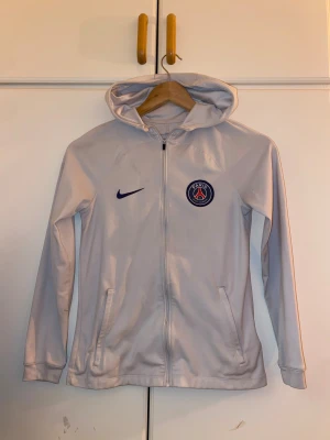 Vit Nike PSG hoodiejacka med dragkedja - Snygg vit hoodiejacka från Nike med PSG-logga på bröstet och Dri-FIT-material. Jackan har hel dragkedja, huva och två fickor framtill. Perfekt för dig som gillar sportig stil och vill representera Paris Saint-Germain.