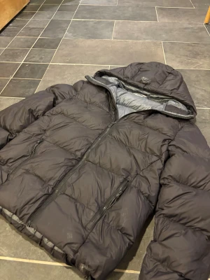 Svart Pufferjacka McKinley - Svart pufferjacka från McKinley med huva och dragkedja framtill. Jackan har två fickor med dragkedja och är vadderad för extra värme. Perfekt för kalla dagar och har en clean, sportig look. Fodret är grått och jackan har en normal passform.