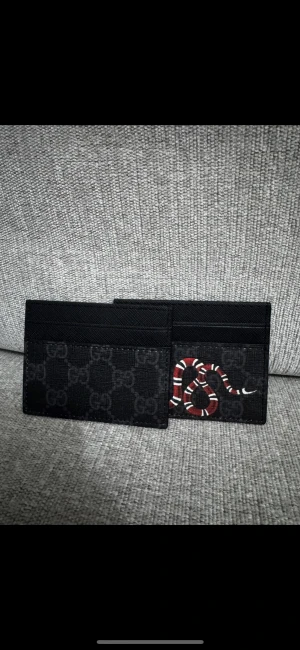 Svart  plånbok med ormprint - Snygg svart plånbok från  med klassiskt GG-mönster och en cool röd och vit ormprint på ena sidan. Slimmad design med flera fack för kort, perfekt att ha i fickan. Materialet är slitstark syntet med exklusiv känsla.