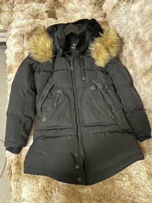 Svart dunjacka med päls från jackan.com - Svart dunjacka från CECIL med fluffig huva och snygga pälsdetaljer i beige/guld. Jackan har flera dragkedjor, stora fickor och ribbade muddar vid ärmslut. Perfekt för kalla dagar med sin quiltade design och långa ärmar.