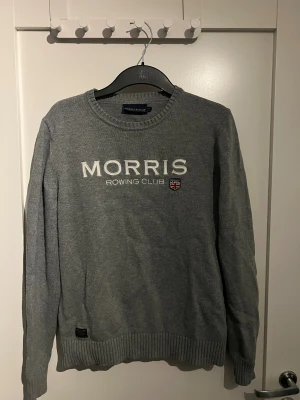 Grå stickad tröja Morris Rowing Club - Grå stickad tröja från Morris Rowing Club med broderad logga och text framtill. Rund hals och ribbade muddar vid ärmslut och nederkant. Perfekt för dig som gillar klassisk stil med sportig touch.