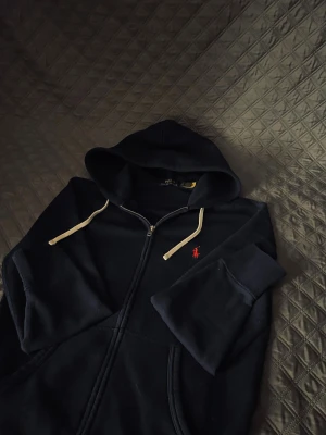 Marinblå hoodie från Ralph Lauren - Snygg marinblå hoodie från Ralph Lauren med dragkedja och huva. Tröjan har vita snören och klassisk röd Polo-logga broderad på bröstet. Tillverkad i mjuk bomull, perfekt för chill dagar. Fickor framtill för extra casual vibe. Den är sparsamt använd, har inte använt mycket, skick 9/10
