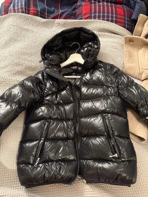 Svart Colmar pufferjacka med huva - Snygg svart pufferjacka från Colmar med glansig finish och huva. Jackan har två dragkedjefickor framtill och tydlig logga på ärmen. Perfekt för kalla dagar och riktigt skön att bära. Stängs med dragkedja hela vägen upp.