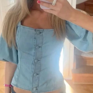 Blå jeans blus med puffärmar - Säljer en snygg ljusblå korsettopp med puffiga korta ärmar och markerad midja. Toppen har knappar framtill och en fyrkantig urringning som ger en trendig vibe. Materialet är i jeans tyg liknade.