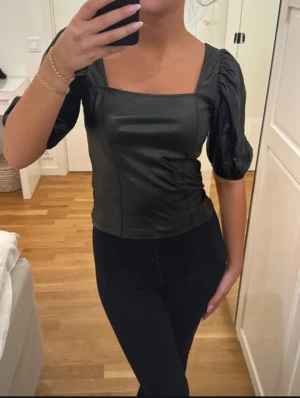 Svart skin topp - Säljer en svart topp med fyrkantig halsringning och puffiga korta ärmar. Toppen har en snygg figurnära passform och är gjord i ett skinnliknande material som ger en cool vibe. Priset går att diskutera 💕💕