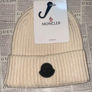 Cream white ribbstickad mössa från Moncler - Snygg vit ribbstickad mössa från Moncler med svart logopatch framtill. Mössan har uppvikt kant och är tillverkad i mjukt material som håller dig varm. Perfekt för dig som vill ha en clean och exklusiv look.