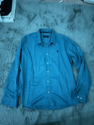 Blå skjorta från Polo Ralph Lauren  - Det står XL men passar mer M . Klassisk design med krage, vita knappar och ikoniska broderade loggan på bröstet. Skjortan är tillverkad i mjuk bomull och har långärmad passform som funkar till många olika stilar.