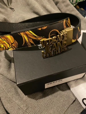  Versace Jeans Couture bälte - Säljer ett sjukt snyggt bälte från Versace Jeans Couture i svart skinn med guldiga barock-inspirerade detaljer och en stor metallspänne med logotyp. Bältet har en lyxig vibe och sticker ut med sitt unika mönster och feta design. Perfekt för dig som vill ha något extra till outfiten.