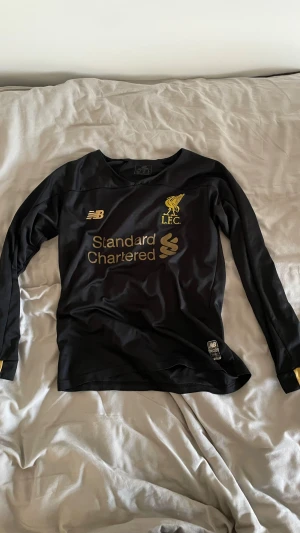 Liverpool målvaktströja A. Becker - Säljer en svart långärmad Liverpool målvaktströja från New Balance med guldiga detaljer och tryck. Framsidan har LFC-logga och Standard Chartered, baksidan har A. Becker och nummer 1. Materialet är lätt och andas, perfekt för fotbollsträning eller match.