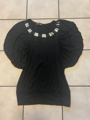 Svart blus med pärldetaljer från K Girls storlek S/M - Svart blus med puffiga korta ärmar och bred mudd nedtill. Runt halsen sitter coola pärldetaljer i svart, vitt och silver som ger extra edge. Materialet är mjukt och stretchigt, perfekt för dig som gillar att sticka ut med din stil.