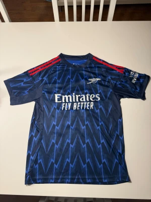 Arsenal bortatröja Gyökeres 14 Adidas - Arsenal bortatröja från Adidas med Gyökeres och nummer 14 på ryggen. Tröjan är mörkblå med blixtmönster i blått, röda axelränder och vita tryck. Tillverkad i lätt och ventilerande polyester, perfekt för match eller träning. Aldrig använd. Ej äkta!