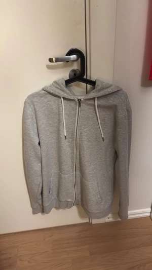 zip-hoodie  - Snygg grå hoodie med dragkedja från 157 i storlek M. Utrustad med huva och vita dragsnören, samt två praktiska fickor framtill. Tillverkad i mjukt och bekvämt sweatshirtmaterial som passar perfekt för chill dagar. Enkel och stilren design.