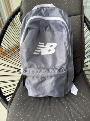 Grå ryggsäck från New Balance - Snygg grå ryggsäck från New Balance med vit logga på framsidan och vita detaljer. Ryggsäck från New Balance i fint skick.  Endast en liten fläck på baksidan, syns knappt (se bild) ! 