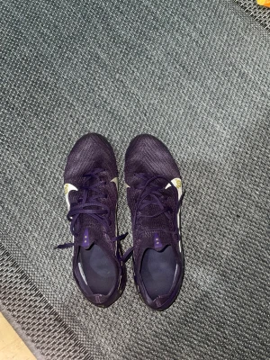 Lila Nike Mercurial fotbollsskor - Säljer ett par Nike Mercurial fotbollsskor i mörklila med guldiga detaljer och snörning. Skorna har en smal och sportig form, mönstrade grafiska inslag i rosa och gult på sidorna samt en platt sula med dobbar för gräsplan. Materialet är syntet och mesh för lätthet och ventilation.