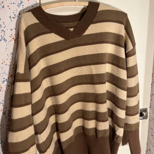 Randig beige och brun tröja - Säljer en stickad tröja med breda bruna och beige ränder. Tröjan har v-ringad hals och långa ärmar med ribbade muddar. Skulle säga storlek M.