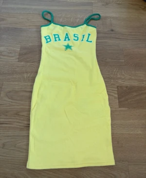 Gul Brasil klänning från FB Sister - Gul kortklänning från FB Sister med gröna detaljer och texten 'BRASIL' framtill. Klänningen har smala axelband och är figurnära i modellen. Perfekt för dig som vill sticka ut med en sportig och färgglad look.OANVÄND!